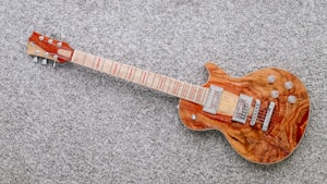 A wooden electric guitar with a natural finish rests on a textured gray carpet. It features a glossy body with intricate wood grain patterns and metallic hardware components such as pickups and control knobs. The guitar neck contains distinct red fret markings.