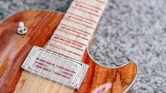 A close-up view of an electric guitar featuring a polished wooden body with a glossy finish. The focus is on the neck and strings, which are positioned over a metallic pickup. The background consists of a textured gray carpet, creating a contrast with the shiny and smooth surface of the guitar.