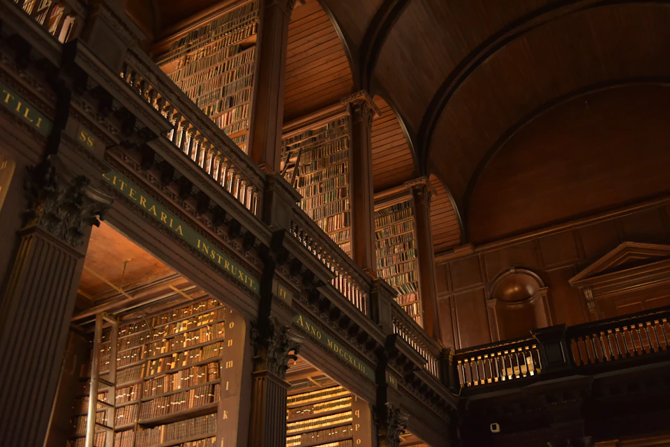 The image depicts a grand, old library with a high ceiling and large wooden beams that support the structure. The walls are lined with numerous bookshelves filled with an array of books, creating a sense of depth and knowledge within the space. A large archway can be seen in the background, adding to the architectural charm of the scene. The lighting is dim, casting long shadows and highlighting the intricate details of the wooden beams and columns that support the ceiling.