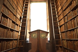A narrow, high-ceilinged library with tall wooden bookshelves filled with numerous brown volumes. Two ladders are positioned on either side of the room, providing access to the upper shelves. A wooden lectern stands in the center with the letters 'P' and 'O' inscribed on it. Light filters in through a large window at the back, illuminating the scene.