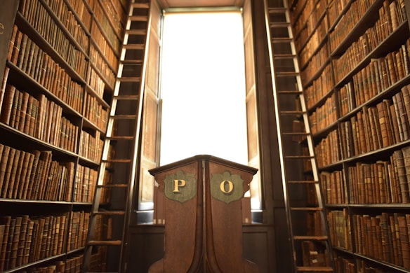 A narrow, high-ceilinged library with tall wooden bookshelves filled with numerous brown volumes. Two ladders are positioned on either side of the room, providing access to the upper shelves. A wooden lectern stands in the center with the letters 'P' and 'O' inscribed on it. Light filters in through a large window at the back, illuminating the scene.