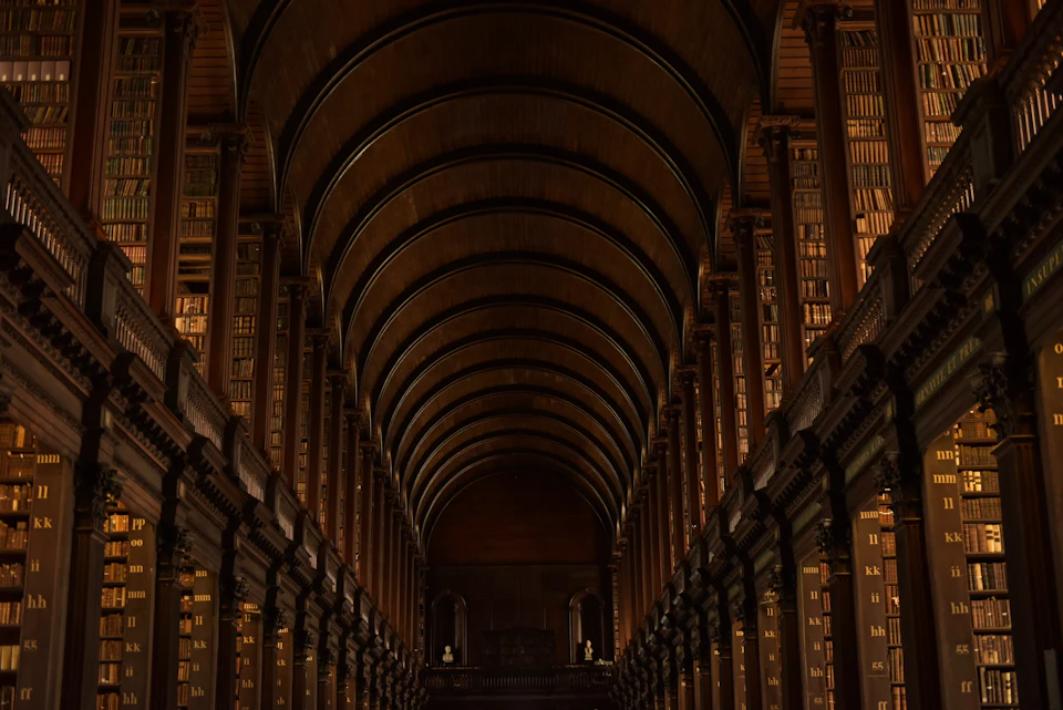 The image depicts a grand, long library with high ceilings and arched windows that allow natural light to filter into the space. The walls are lined with rows upon rows of bookshelves filled with books of various sizes and colors, creating an atmosphere of knowledge and learning. The perspective of the photo is from the entrance of the library, looking down a hallway that extends towards the end of the building.