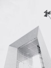 A large, modern, geometric building with a noticeable rectangular arch structure, constructed with a grid pattern. The angle captures the underside and the towering sides of the structure. A minimalist design complements the structure, which includes industrial elements such as grids and panels. In the top right corner, there is a unique, abstract sculpture mounted on a pole, adding an artistic touch to the architectural scene.