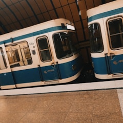 Two subway trains with blue and cream exteriors are stationed on parallel tracks inside an underground train station. The train station features a brown tiled floor and a curved ceiling with large windows.