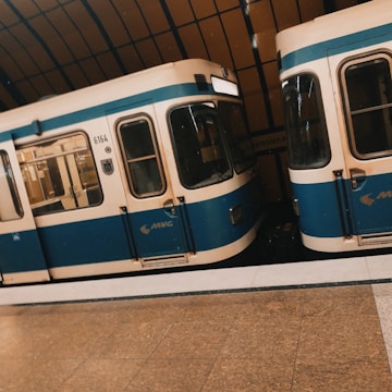 Two subway trains with blue and cream exteriors are stationed on parallel tracks inside an underground train station. The train station features a brown tiled floor and a curved ceiling with large windows.