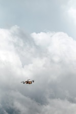 A drone is flying in a cloudy sky, surrounded by large, thick clouds that dominate the background. The drone is small in size compared to the vast expanse of clouds, suggesting it is at a considerable height.