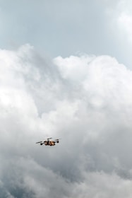 A drone is flying in a cloudy sky, surrounded by large, thick clouds that dominate the background. The drone is small in size compared to the vast expanse of clouds, suggesting it is at a considerable height.
