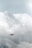 A drone is flying in a cloudy sky, surrounded by large, thick clouds that dominate the background. The drone is small in size compared to the vast expanse of clouds, suggesting it is at a considerable height.