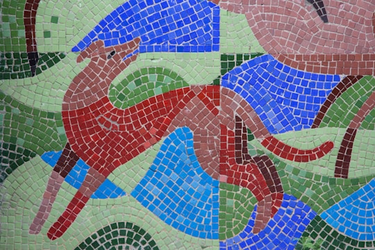 A colorful mosaic depicts a stylized dog or similar animal in motion, formed by small square tiles. The animal is primarily red and brown with a playful expression, surrounded by abstract shapes in blue, green, and light brown. The background is a combination of blue and green wavy patterns.