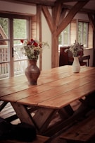 A wooden dining table with a rustic finish is set in a room with large windows. Two ceramic vases containing red, blue, and white flowers are placed on top of the table. The room features exposed wooden beams, giving it a cozy, natural feel. Soft light filters through the windows, highlighting the texture of the wood.
