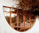 A round window set within a white brick wall with reflective glass panes that mirror surrounding trees. The wooden frame of the window contrasts with the white bricks, and branches with green leaves partially cover the upper right portion of the window.