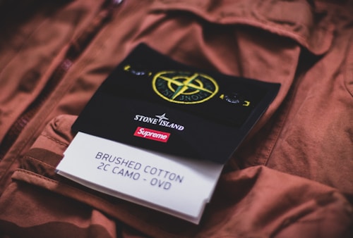 A close-up view of a garment featuring a tag with the Stone Island and Supreme logos. The tag includes details about the material, stating it is made of brushed cotton in a 2C camo pattern. The background shows the fabric of a brown jacket or coat.