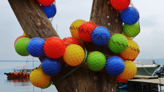 A collection of colorful spherical floats, covered in netting, are wrapped around a large wooden structure. In the background, a calm body of water is visible along with boats docked at a pier. The sky is overcast, providing a muted backdrop to the vibrant colors of the floats.