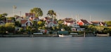 A serene coastal village with multiple red wooden houses featuring white trims, surrounded by lush greenery and trees. Several Swedish flags are visible, fluttering gently in the breeze. A calm body of water occupies the foreground with a small dock and a white wooden boat moored next to it.