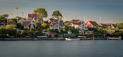 A serene coastal village with multiple red wooden houses featuring white trims, surrounded by lush greenery and trees. Several Swedish flags are visible, fluttering gently in the breeze. A calm body of water occupies the foreground with a small dock and a white wooden boat moored next to it.