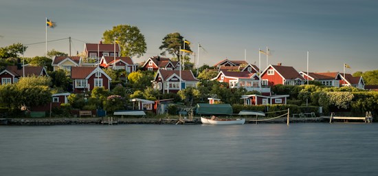 A serene coastal village with multiple red wooden houses featuring white trims, surrounded by lush greenery and trees. Several Swedish flags are visible, fluttering gently in the breeze. A calm body of water occupies the foreground with a small dock and a white wooden boat moored next to it.