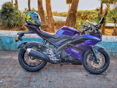 A sleek blue and purple Yamaha motorcycle is parked on a brick pavement next to a bright blue wall with a mosaic detail. A colorful motorcycle helmet rests on the seat. In the background, there are tall trees and some lush green foliage, with a glimpse of water and more greenery in the distance.