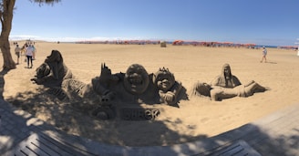 Sand sculptures of characters from a popular animated movie, including a large smiling figure and others with distinct facial expressions. The sculptures are detailed and surrounded by tourists. The beach is expansive with clear skies and orange umbrellas in the distance.
