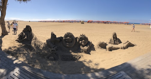 Sand sculptures of characters from a popular animated movie, including a large smiling figure and others with distinct facial expressions. The sculptures are detailed and surrounded by tourists. The beach is expansive with clear skies and orange umbrellas in the distance.