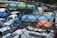 A cluttered junkyard filled with a variety of old vehicles, including buses, trucks, and vans, tightly packed together. The vehicles are in various states of disrepair, with some missing parts and others rusted. The scene is framed by a wire mesh fence, suggesting restricted access.