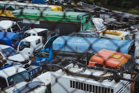 A cluttered junkyard filled with a variety of old vehicles, including buses, trucks, and vans, tightly packed together. The vehicles are in various states of disrepair, with some missing parts and others rusted. The scene is framed by a wire mesh fence, suggesting restricted access.