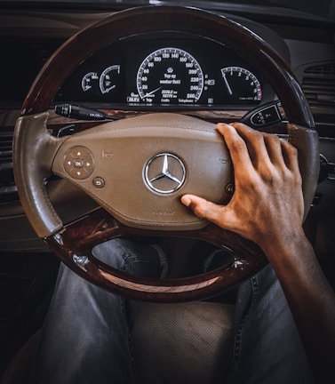 A person is gripping a Mercedes-Benz steering wheel with one hand, with the dashboard and instrument panel visible in the background displaying various gauges and notifications.