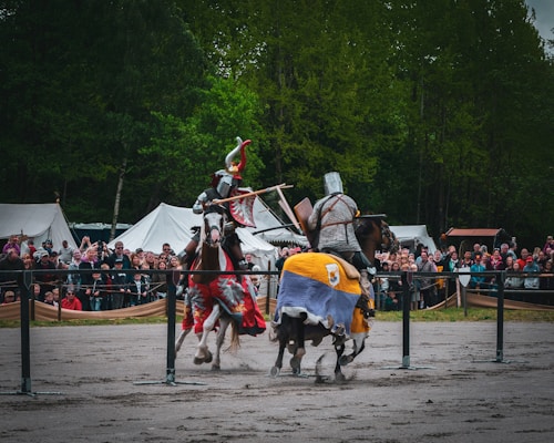 Two knights clad in traditional armor are jousting on horseback in a medieval-style event. Both horses are adorned with colorful caparisons, and the knights hold wooden lances aimed at each other. The background features white tents and a large crowd of spectators eagerly watching the event.