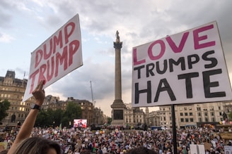 A large crowd gathered in an urban square, holding protest signs reading 'Dump Trump' and 'Love Trumps Hate'. The scene is set against a backdrop of historic buildings and a tall monument. The overcast sky adds to the atmosphere of the event.