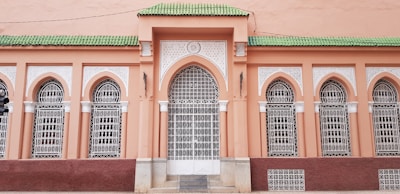 A facade with ornate architectural details features arched windows fitted with intricate white wrought iron grilles. The wall is painted in a warm peach hue, while the roofline is accented by vibrant green tiles.