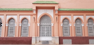 A facade with ornate architectural details features arched windows fitted with intricate white wrought iron grilles. The wall is painted in a warm peach hue, while the roofline is accented by vibrant green tiles.