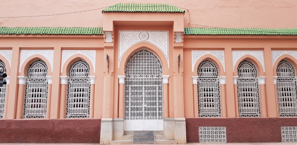 A facade with ornate architectural details features arched windows fitted with intricate white wrought iron grilles. The wall is painted in a warm peach hue, while the roofline is accented by vibrant green tiles.
