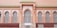 A facade with ornate architectural details features arched windows fitted with intricate white wrought iron grilles. The wall is painted in a warm peach hue, while the roofline is accented by vibrant green tiles.