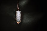 A vertically oriented crystal pendant encased in a wire wrapping, suspended from a chain. The crystal appears to be smooth and polished, with a cylindrical shape. The wire wrapping is intricately designed to hold the crystal securely, and the lighting casts a soft glow that highlights the textures.