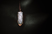 A vertically oriented crystal pendant encased in a wire wrapping, suspended from a chain. The crystal appears to be smooth and polished, with a cylindrical shape. The wire wrapping is intricately designed to hold the crystal securely, and the lighting casts a soft glow that highlights the textures.