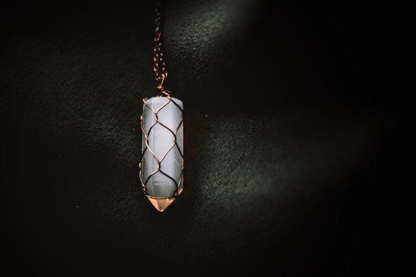 A vertically oriented crystal pendant encased in a wire wrapping, suspended from a chain. The crystal appears to be smooth and polished, with a cylindrical shape. The wire wrapping is intricately designed to hold the crystal securely, and the lighting casts a soft glow that highlights the textures.