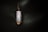 A vertically oriented crystal pendant encased in a wire wrapping, suspended from a chain. The crystal appears to be smooth and polished, with a cylindrical shape. The wire wrapping is intricately designed to hold the crystal securely, and the lighting casts a soft glow that highlights the textures.