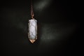 A vertically oriented crystal pendant encased in a wire wrapping, suspended from a chain. The crystal appears to be smooth and polished, with a cylindrical shape. The wire wrapping is intricately designed to hold the crystal securely, and the lighting casts a soft glow that highlights the textures.
