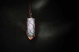 A vertically oriented crystal pendant encased in a wire wrapping, suspended from a chain. The crystal appears to be smooth and polished, with a cylindrical shape. The wire wrapping is intricately designed to hold the crystal securely, and the lighting casts a soft glow that highlights the textures.