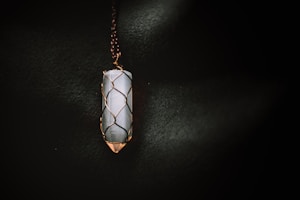 A vertically oriented crystal pendant encased in a wire wrapping, suspended from a chain. The crystal appears to be smooth and polished, with a cylindrical shape. The wire wrapping is intricately designed to hold the crystal securely, and the lighting casts a soft glow that highlights the textures.