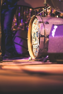 A drum with a colorful, intricate skull design is prominently displayed. The surrounding area is dimly lit with purple lighting, creating an atmospheric and moody setting. Part of a drum set is visible, and nearby, the leg of a person wearing jeans can be seen.