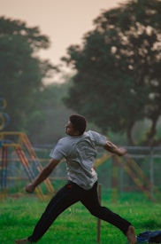 A person is captured mid-action, appearing to be in a dynamic pose as if throwing or swinging something. The scene takes place outdoors with a backdrop of trees and playground equipment in a park-like setting. The lighting suggests it might be late afternoon or early evening, giving a soft, natural light to the scene.