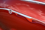 A close-up of a shiny, red car with a metallic emblem and decorative flame-like designs alongside grooves. The reflections on the surface suggest a smooth and polished finish.