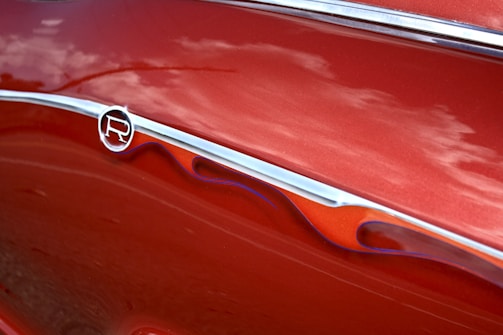 A close-up of a shiny, red car with a metallic emblem and decorative flame-like designs alongside grooves. The reflections on the surface suggest a smooth and polished finish.
