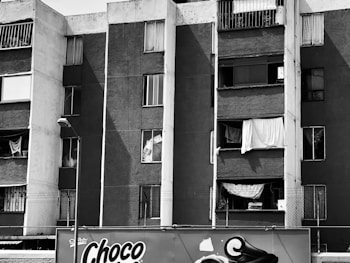 A multi-story residential building with a worn exterior. The facade features several windows with missing or makeshift coverings, such as sheets and curtains. Below, there is an advertisement for a product named 'Choco' on a billboard.