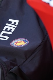 A close-up view of a bag featuring a logo with a lion and the words 'ROYALS FC'. The bag is primarily dark in color with the word 'FIELD' printed on it in white. A bright red colored background is partially visible.