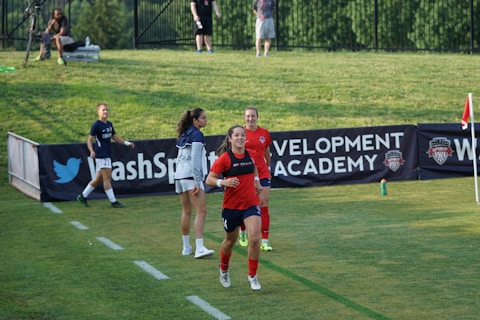 A woman in a red soccer jersey runs on a grassy field near a corner flag, smiling as she moves with energy and purpose. Behind her, two other women in sports attire stand on the field, and a few spectators are visible in the background. A large banner is displayed in the background with sports-related text, adding context to the setting as a soccer game or training session.