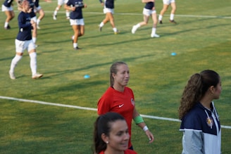 A group of female soccer players is on a field. One player in a red jersey walks in the foreground with a slight smile. Other players in dark blue jerseys are in the background, some moving or practicing. The grass is vivid green with small blue cones placed on it.