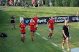 A group of female soccer players wearing red jerseys are walking across a grassy field. They are accompanied by a woman in a black shirt and shorts who appears to be focused on something ahead. In the background, there are several people observing from behind a barrier displaying banners. The setting suggests a sports event or training session.