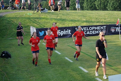 A group of female soccer players wearing red jerseys are walking across a grassy field. They are accompanied by a woman in a black shirt and shorts who appears to be focused on something ahead. In the background, there are several people observing from behind a barrier displaying banners. The setting suggests a sports event or training session.