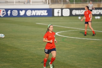 Two female soccer players are on a soccer field wearing red and dark blue uniforms. One player is in the foreground running with a green headband and the number 31 on her shorts, while the other player is in the background. The field has markings and soccer balls are scattered around. Advertising banners with social media icons and text are visible in the background.